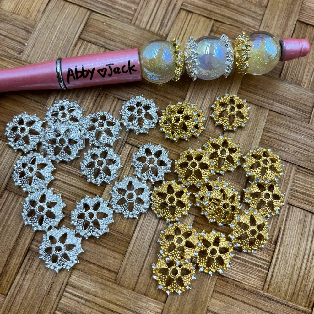 Silver and gold filigree flower-shaped rhinestone bead caps displayed on wicker surface next to a beadable pen, used for DIY jewelry making and beadable pens.