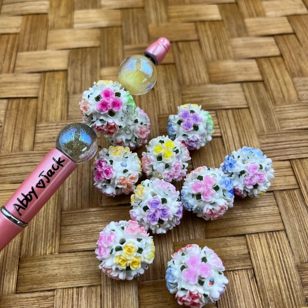 Colorful floral bouquet ball beads covered in tiny pastel mini roses in pink, yellow, purple, and blue, displayed next to a pink beadable pen on a woven bamboo surface.