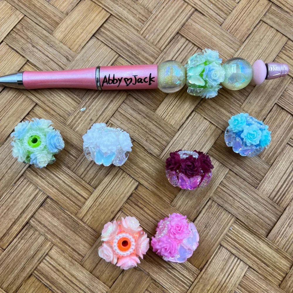 Assorted iridescent resin fancy beads with floral rose clusters in blue, pink, white, purple, and green, displayed alongside a personalized pink glitter beadable pen on a woven surface.