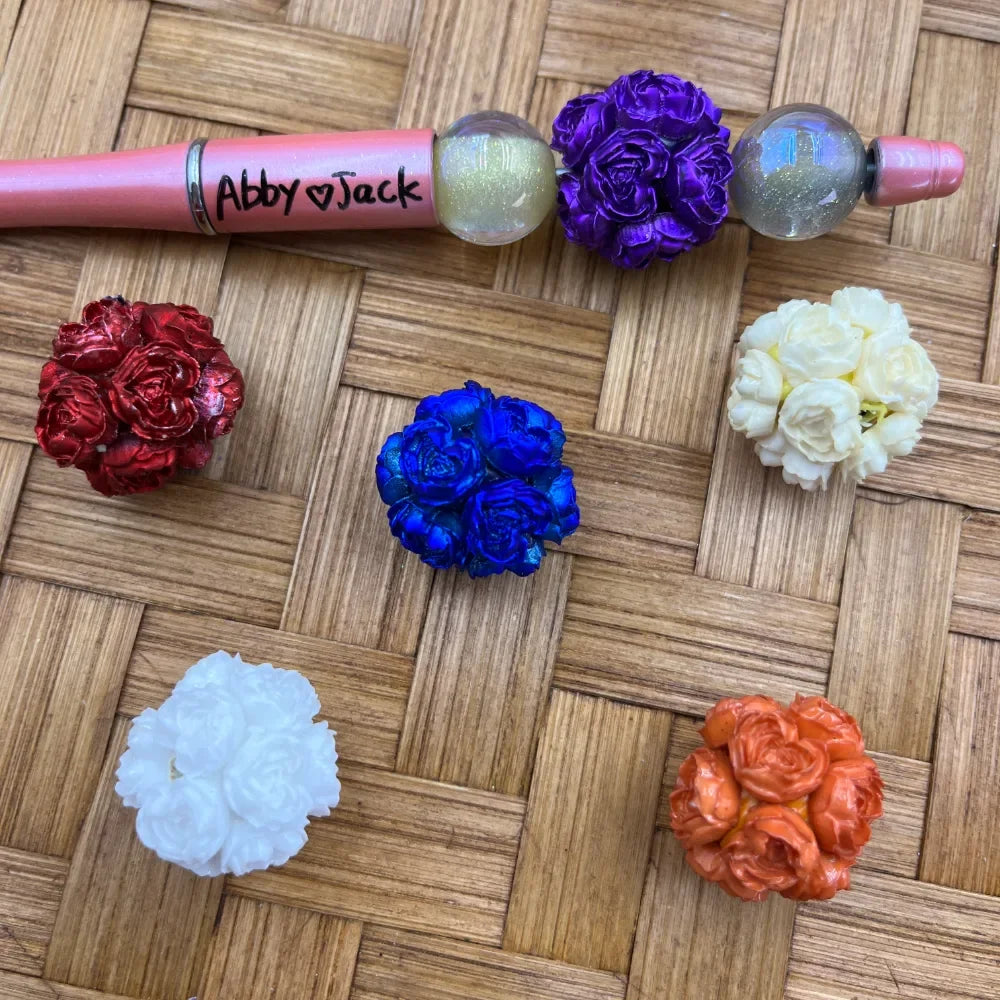 Assorted floral cluster rose ball fancy beads in red, royal blue, purple, white, cream, and orange displayed on a woven bamboo surface next to a pink beadable pen labeled Abby and Jack.