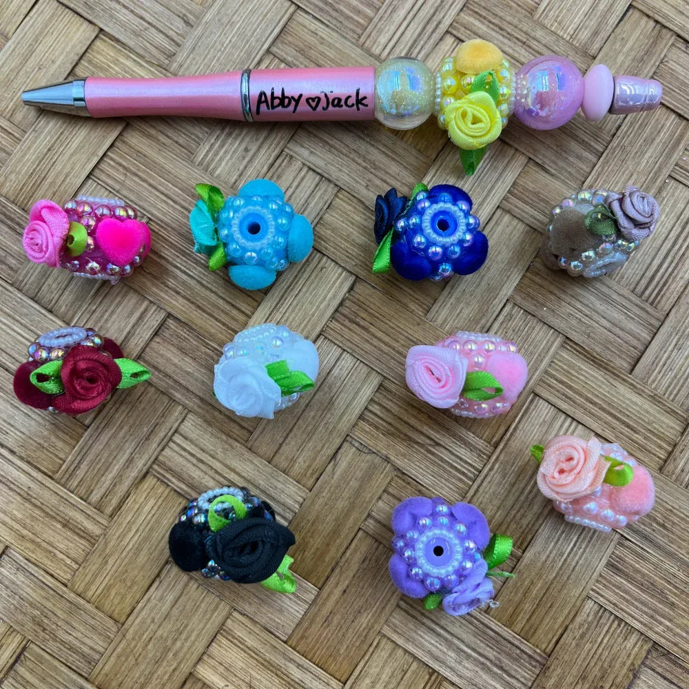 Assorted colorful floral fancy beads with pearl accents and satin ribbon roses in pink, blue, purple, white, black, and red, displayed on a woven surface next to a decorated beadable pen.