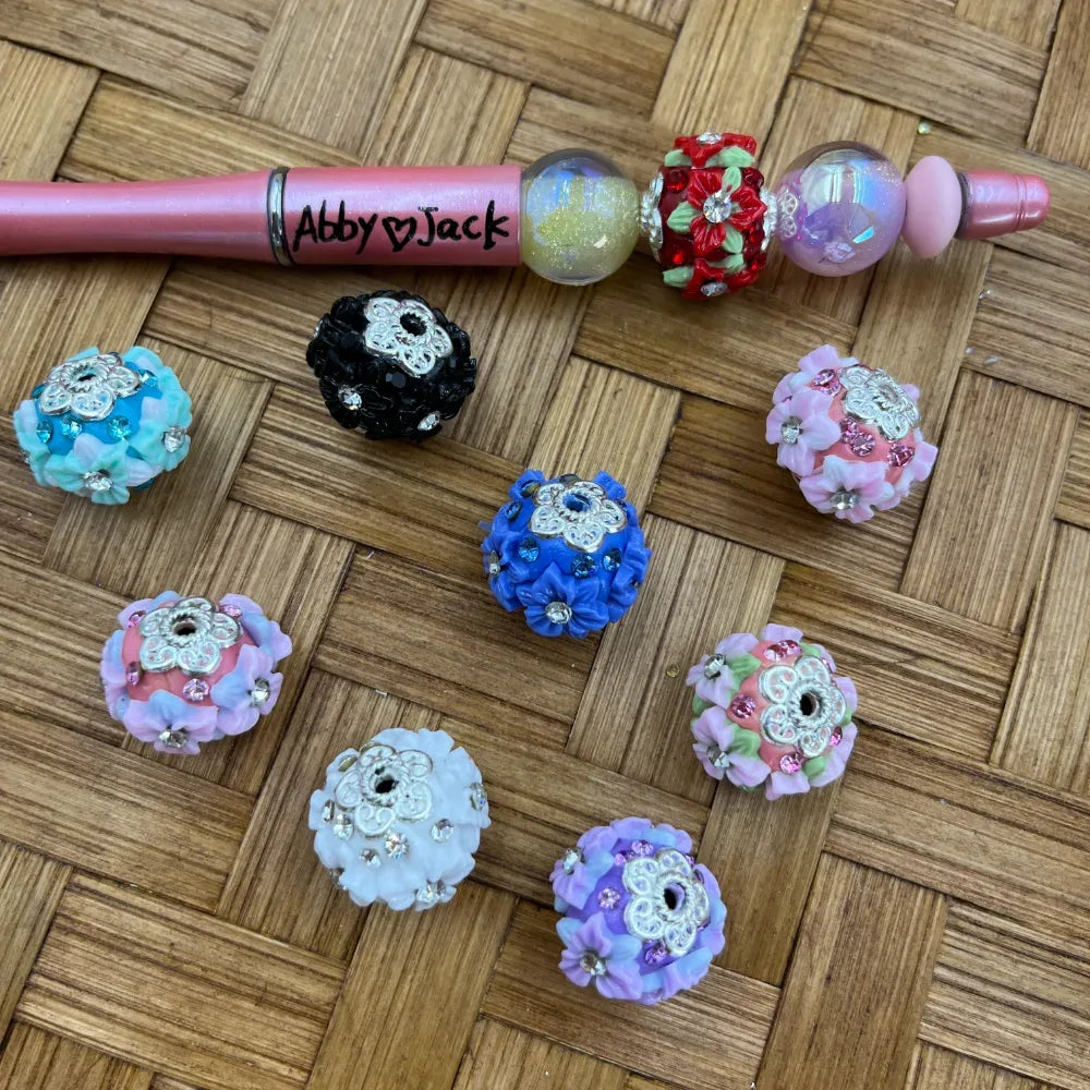 Assorted floral rhinestone clay fancy beads in multiple colors including blue, pink, white, lavender, black, and red arranged on a wicker surface next to a pink beadable pen labeled Abby Jack.