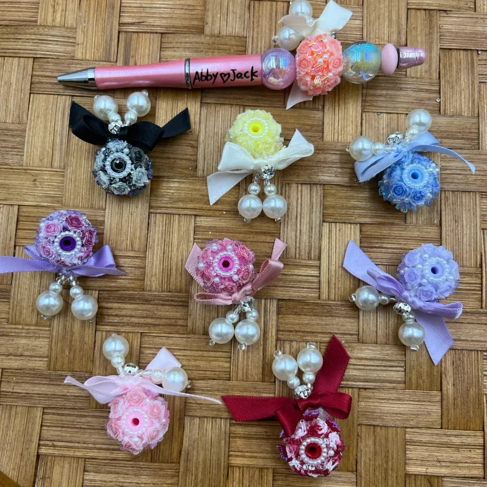 Decorative floral rose ball fancy beads with white pearl dangles and ribbon bows in assorted colors including pink, lavender, blue, black, red, and yellow, displayed on a woven surface alongside a personalized beadable pen labeled Abby and Jack.