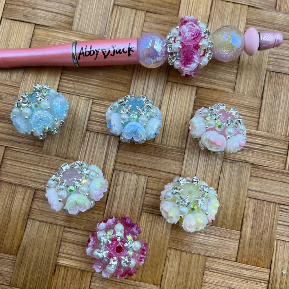 Assorted floral rose fancy beads with rhinestones and pearl accents in pink, white, and blue colors, displayed on a woven bamboo surface next to a decorated beadable pen branded Abby and Jack.