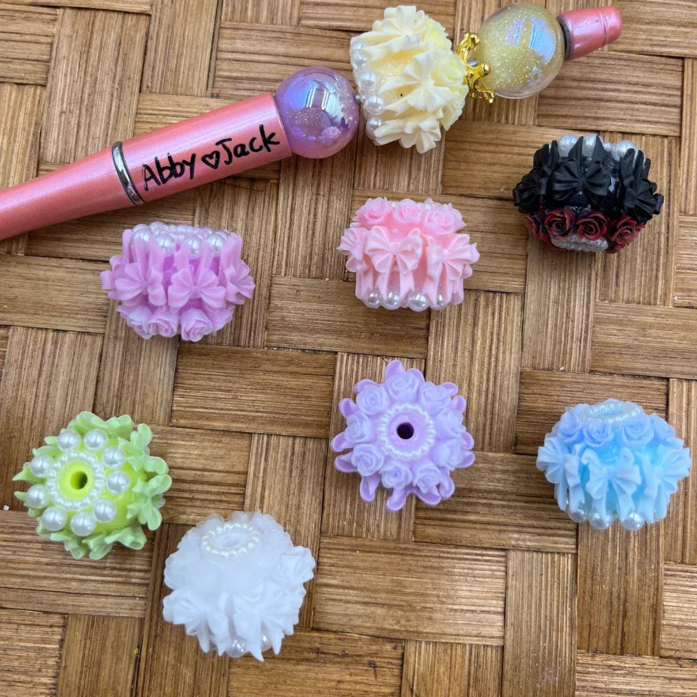 Assorted floral rose ruffle resin fancy beads in pink, purple, white, blue, green, lavender, and black with pearl accents, displayed on a wicker surface alongside a decorated beadable pen labeled Abby and Jack.