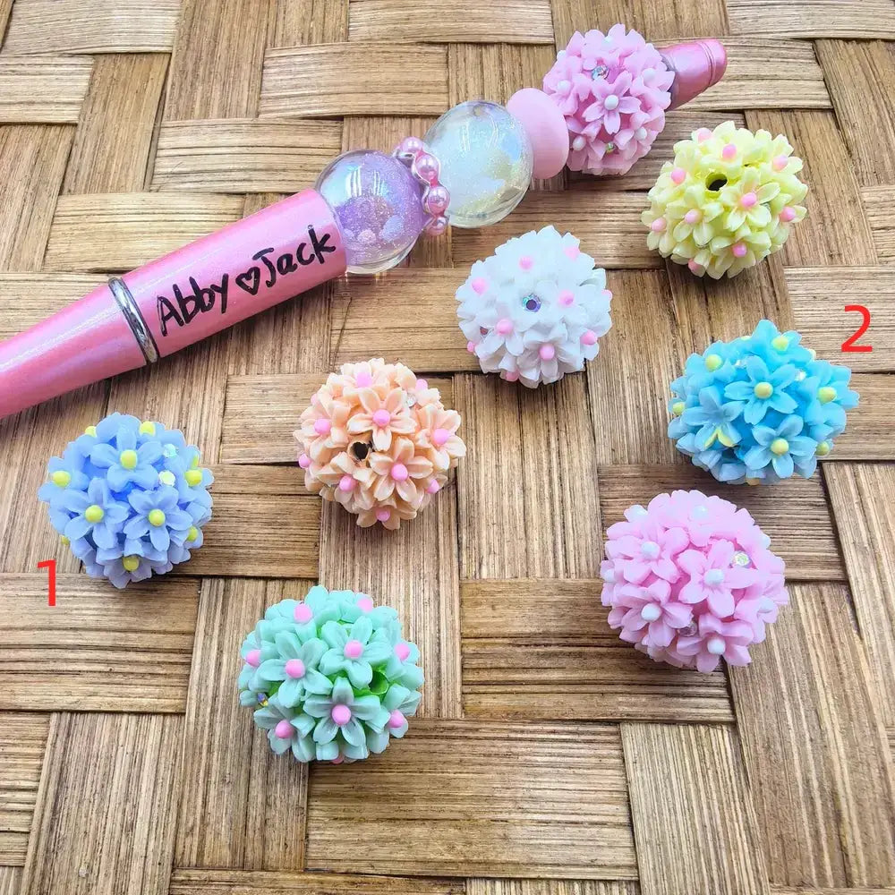 Assorted pastel flower cluster ball resin fancy beads in blue, peach, mint, white, yellow, light blue, and pink, displayed next to a decorative pink beadable pen on a woven surface.