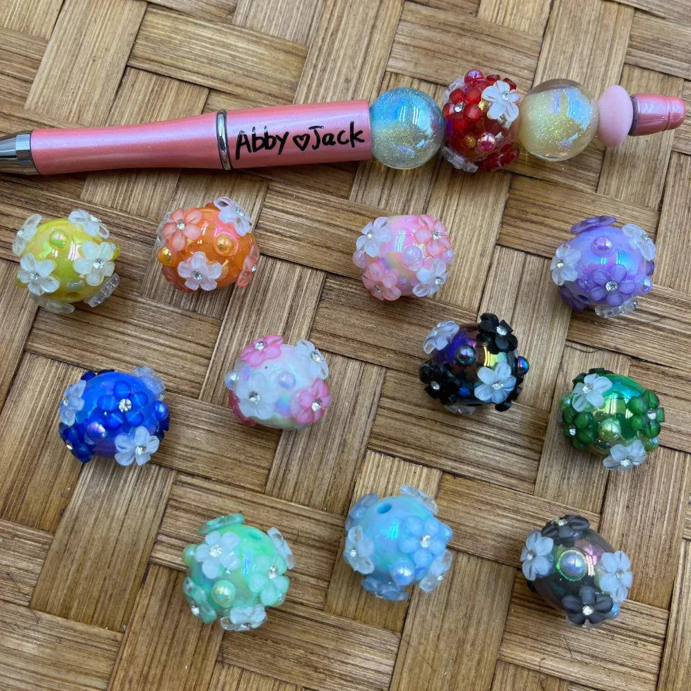 Assorted colorful flower cluster chunky bubblegum beads in yellow, orange, pink, purple, blue, black, green, teal, and gray with rhinestone accents, displayed on a woven bamboo surface next to a beadable pen labeled Abby and Jack.