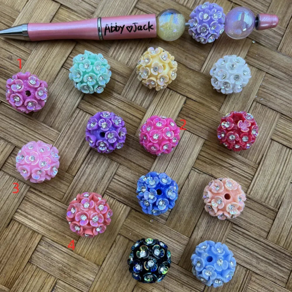 Assorted flower cluster resin beads with rhinestone centers in pink, mint, yellow, purple, red, blue, peach, black, and white, displayed on a woven surface next to a beadable pen.