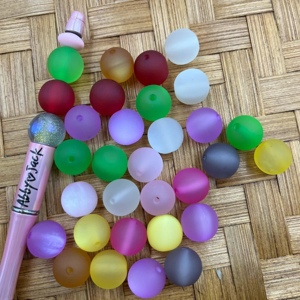 Assorted frosted matte translucent acrylic beads in multiple colors including green, red, purple, yellow, gray, white, and pink displayed on a wicker surface next to a pink beadable pen.