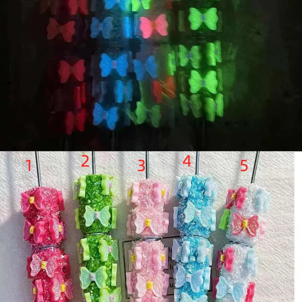 Five colorful glow-in-the-dark butterfly bow rhinestone beadable pens in pink, green, light pink, blue, and light blue, shown glowing in the dark above their daytime appearance.