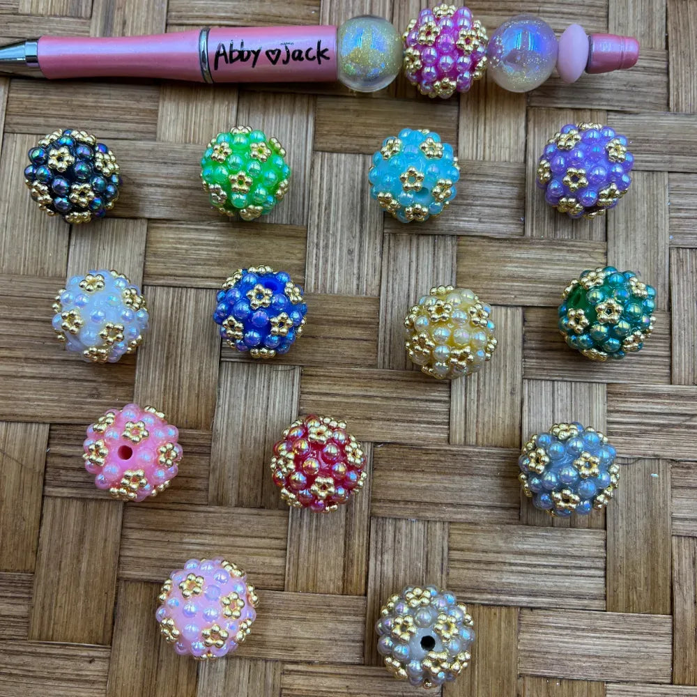 Assorted iridescent gold flower cluster berry beads in 12 colors including black, green, blue, pink, red, purple, teal, and white, displayed on woven bamboo surface next to a beadable pen.