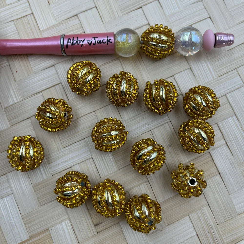 Gold oval fancy beads encrusted with gold rhinestones and crystal accents, displayed on a woven surface next to a pink beadable pen decorated with matching gold rhinestone beads.