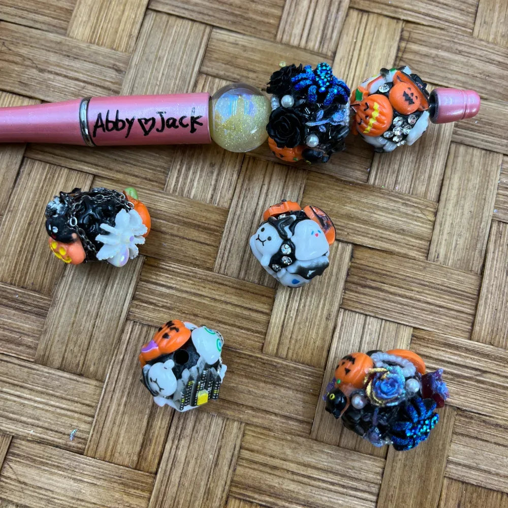 Halloween themed chunky fancy beads featuring jack-o-lantern pumpkins, white ghosts, black roses, blue glitter flowers, and rhinestone accents displayed next to a pink beadable pen labeled Abby Jack on a woven rattan surface.