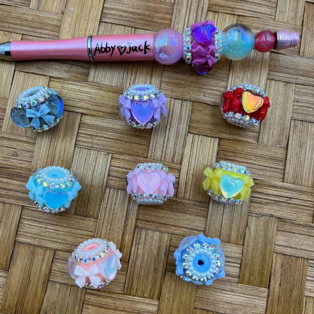 Colorful fancy beads featuring heart centers, flower petal details, rhinestone and pearl trim in blue, purple, red, pink, yellow and teal, displayed on woven bamboo surface alongside a decorated beadable pen labeled Abby Jack.