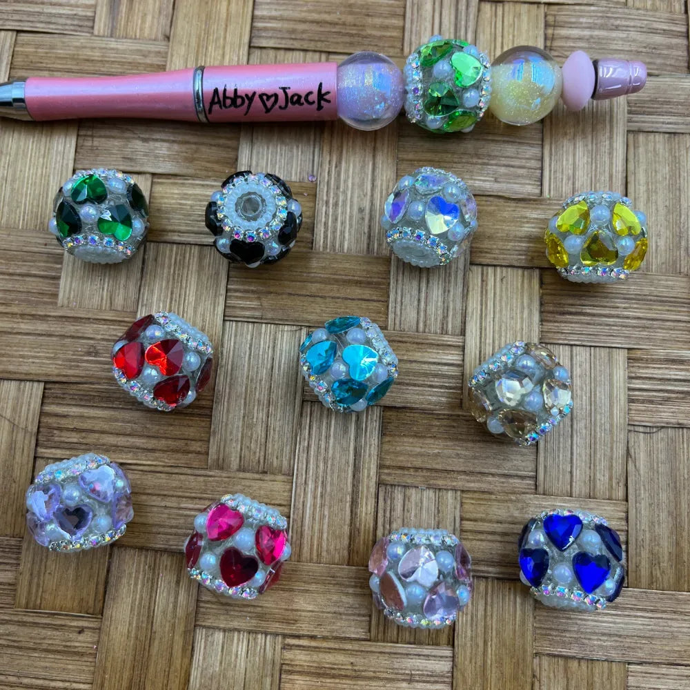 Assorted heart-shaped rhinestone fancy beads in multiple colors including red, blue, pink, green, yellow, teal, and clear crystal, with pearl and AB crystal accents, displayed on a woven surface next to a pink beadable pen labeled Abby and Jack.