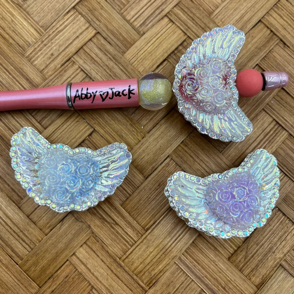 Holographic iridescent angel wing heart beads with rose embossed center and rhinestone border, displayed next to beadable pens on wicker surface