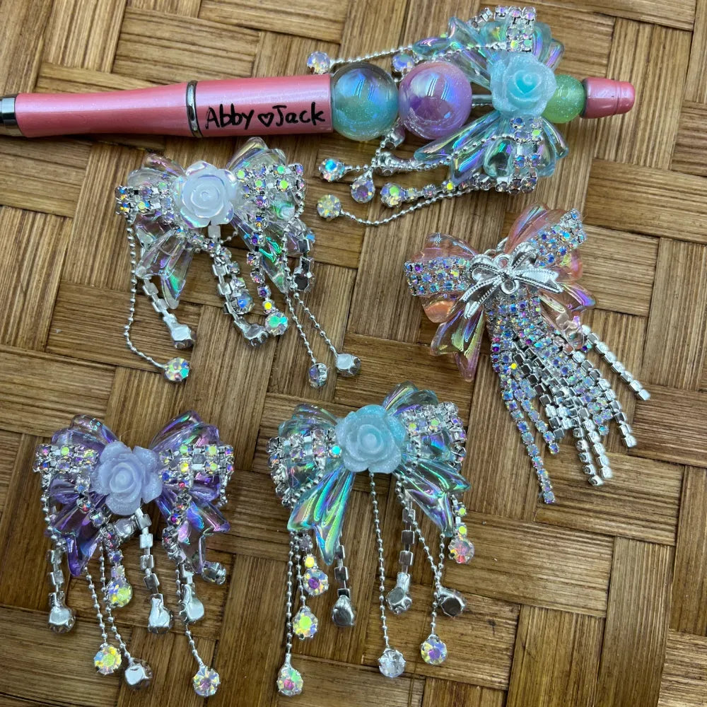 Holographic iridescent bow-shaped fancy beads with rhinestone tassel chains, rose flower accents, and aurora borealis crystal dangles displayed on a woven surface alongside a decorated beadable pen.