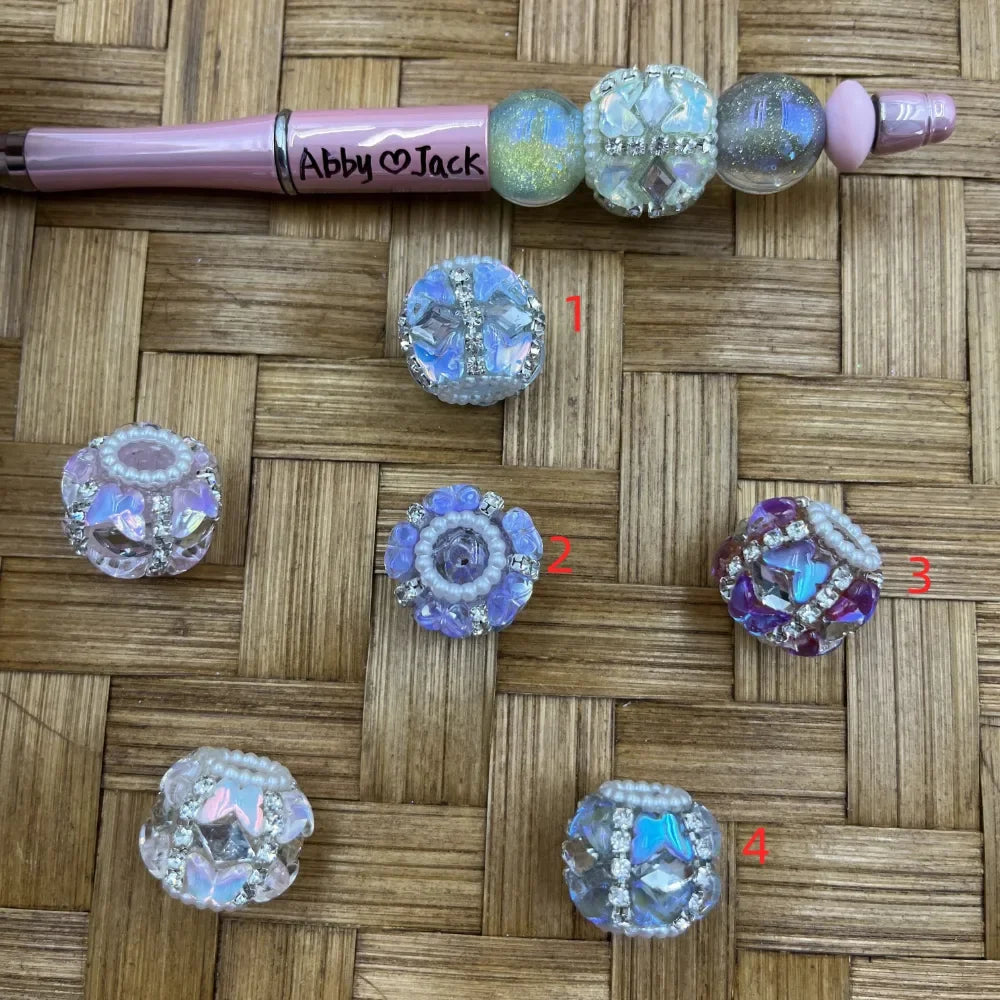 Assorted iridescent AB crystal rhinestone fancy beads in blue, purple, and clear with bling detailing, displayed on wicker surface next to a pink beadable pen labeled Abby and Jack, numbered 1 through 4 showing different bead styles.