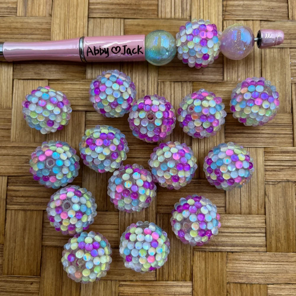 Iridescent pastel rainbow berry cluster acrylic fancy beads displayed on woven surface next to a pink beadable pen labeled Abby heart Jack, showing multicolor pink blue yellow holographic bubble texture beads.