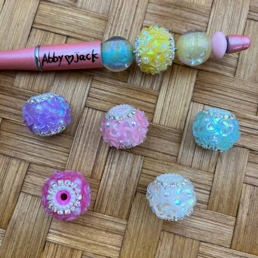 Pastel iridescent bubble textured resin beads with rhinestone bands in purple, pink, teal, yellow, and white, displayed on a woven surface next to a beadable pen labeled Abby and Jack.