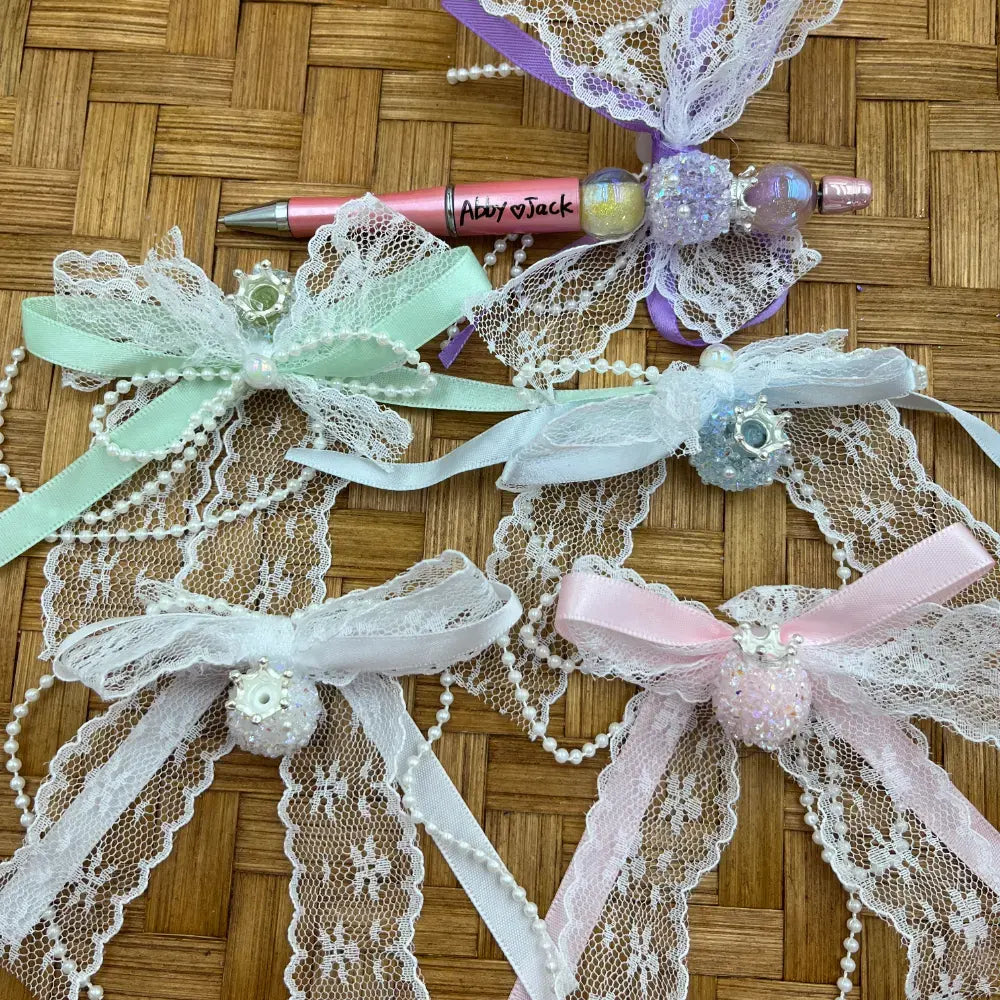 Decorative fancy beads with white lace bows, satin ribbons in mint, lavender, white, and pink, pearl bead chains, and sparkly iridescent center beads displayed on wicker surface alongside a personalized beadable pen.