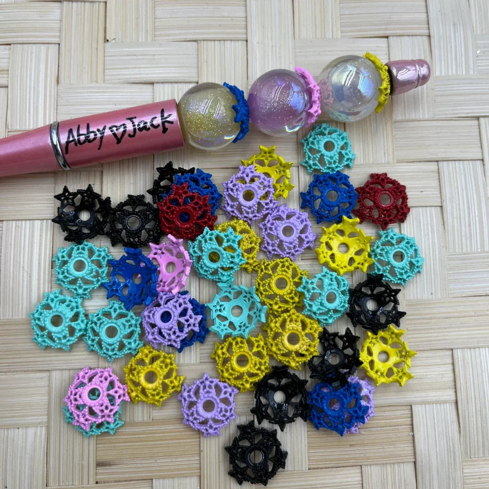 Assorted colorful lace flower bead caps in black, teal, yellow, pink, blue, red, purple, and lavender displayed on a wicker surface next to a pink beadable pen with globe beads from DIY Mini Planet.