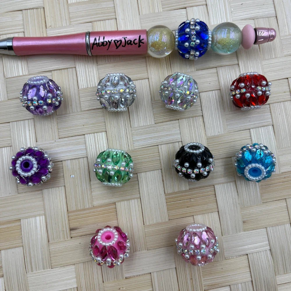 Assorted large acrylic fancy beads with faceted rhinestones and pearl accents in purple, clear, green, black, blue, red, and pink beside a pink beadable pen