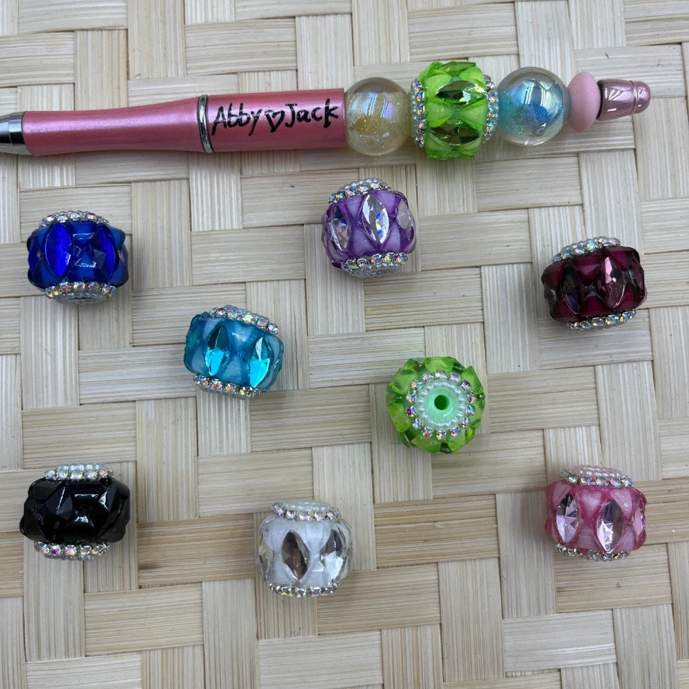 Assorted large hole faceted acrylic fancy beads with rhinestone trim in blue, purple, burgundy, aqua, green, black, white, and pink beside a pink beadable pen.