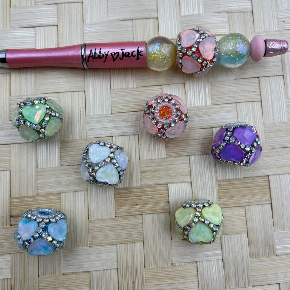 Six pastel heart rhinestone large-hole fancy beads with a pink beadable pen on a woven background