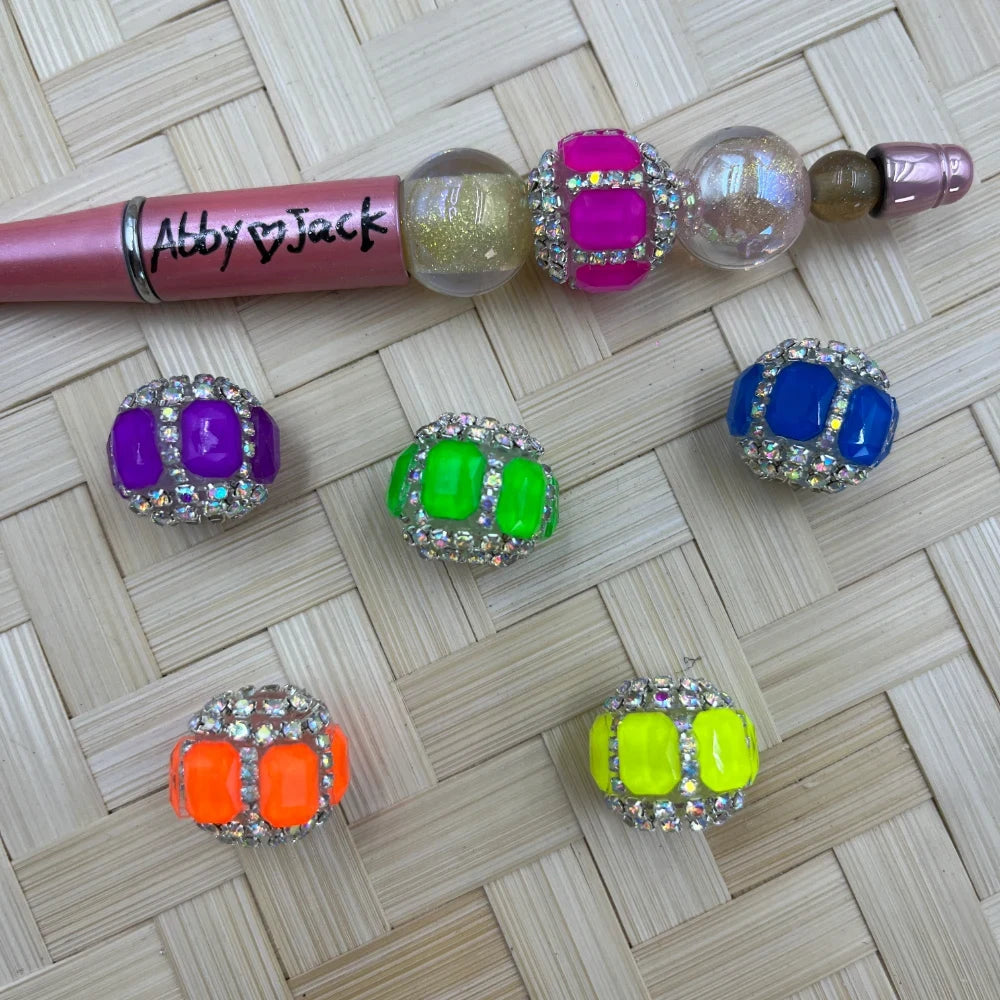 Assorted neon rhinestone square gem fancy beads in purple, neon green, blue, orange, and yellow with crystal rhinestone borders, displayed alongside a beadable pen on a wicker surface.