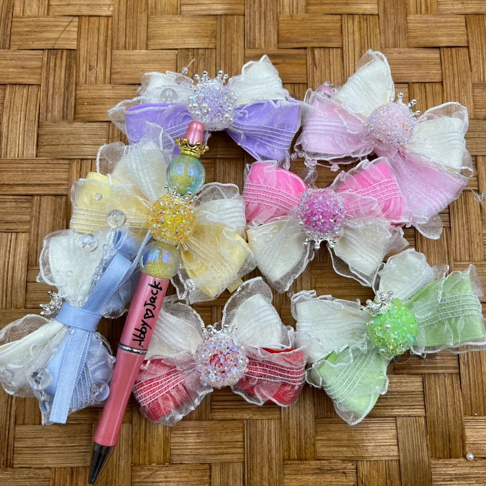 Decorative beadable pens adorned with pastel organza ruffle bows and sparkly crown rhinestone center beads in purple, pink, yellow, blue, red, and green on a wicker background from DIY Mini Planet.