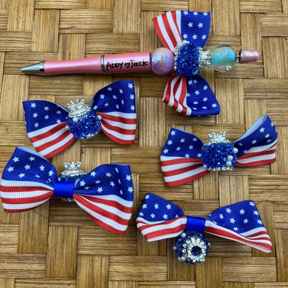 Patriotic American flag ribbon bow fancy beads with blue rhinestone and silver crown embellishments displayed on woven surface alongside a pink beadable pen, ideal for 4th of July DIY crafts.