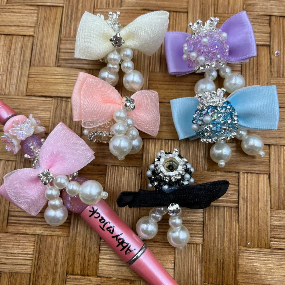 Assorted pastel pearl bow and rhinestone crown fancy beads in white, pink, lavender, blue, and black displayed on woven bamboo surface, perfect for beadable pens and DIY jewelry making at DIY Mini Planet.