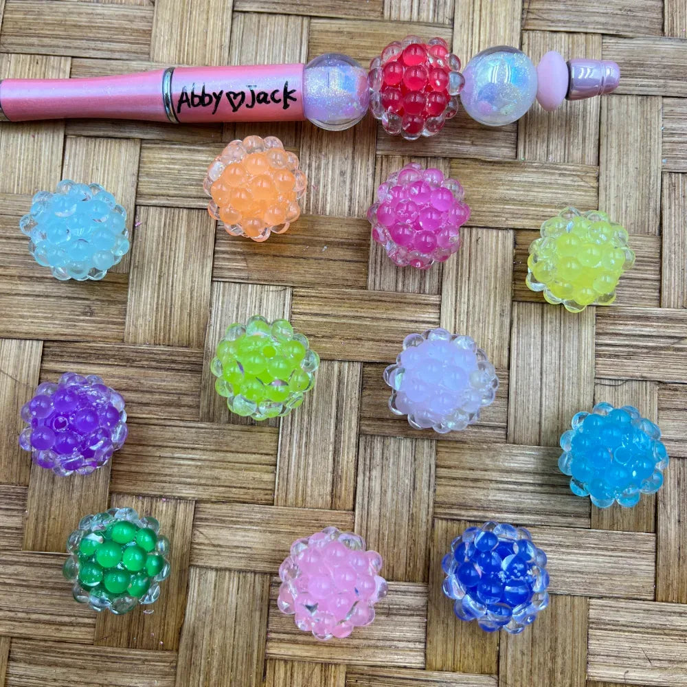 Assorted colorful raspberry bubble cluster acrylic fancy beads in pink, red, orange, green, purple, blue, yellow, and white, displayed on a woven surface next to a pink beadable pen labeled Abby and Jack.