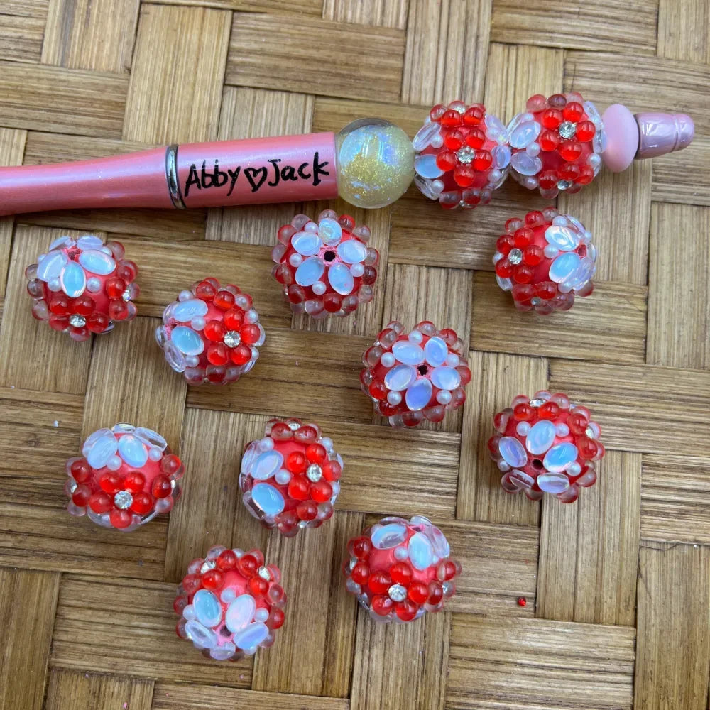 Red chunky beads with white opal flower petals and rhinestone centers, displayed on wicker surface alongside a pink beadable pen labeled Abby Jack