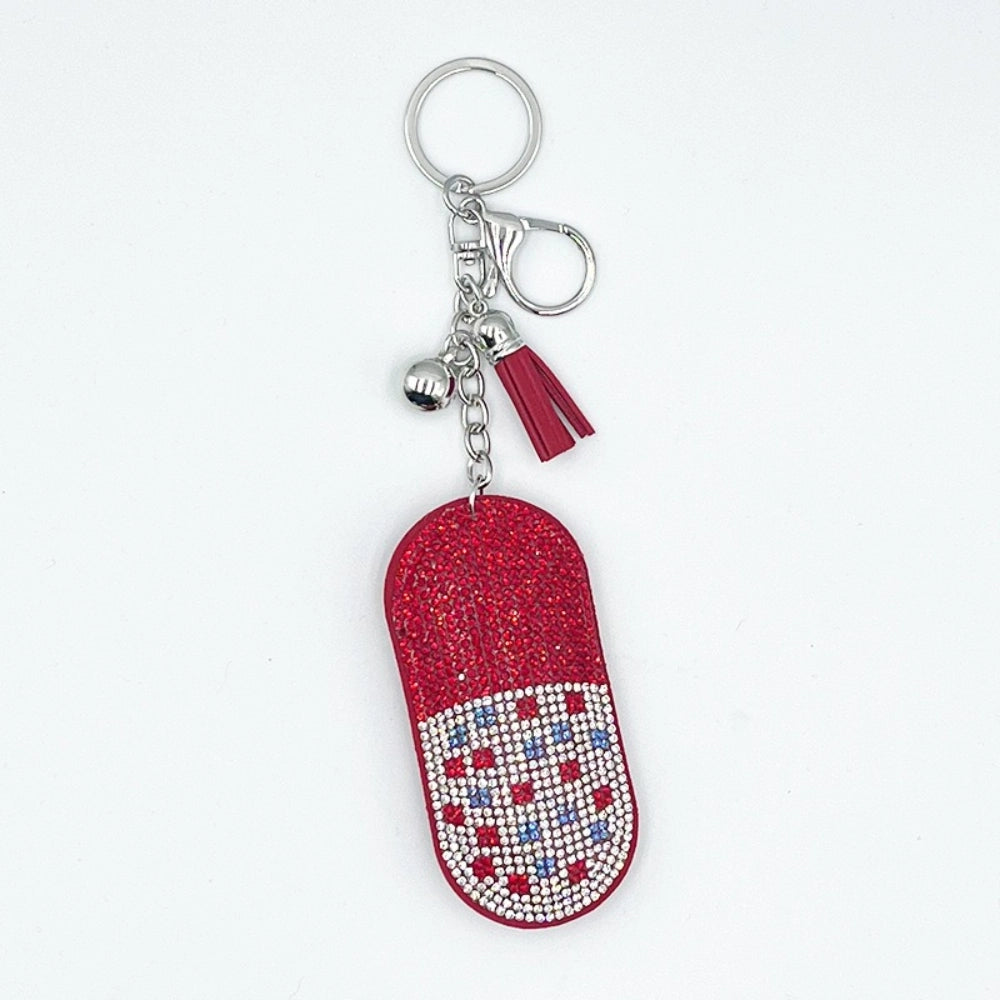 Red glitter rhinestone pill capsule shaped keychain charm with silver bell, red tassel, and crystal bead embellishments on white background.