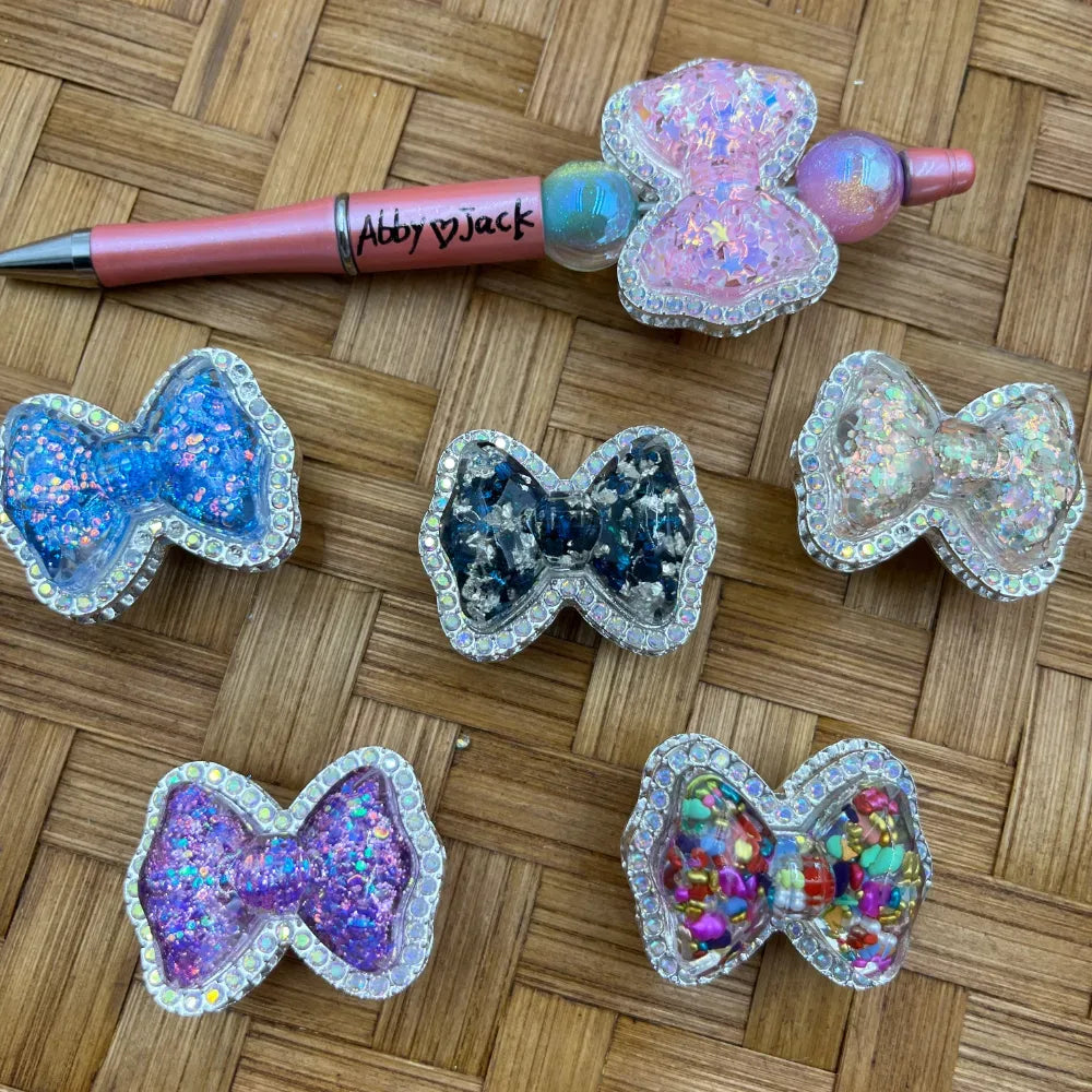 Assorted glitter-filled resin bow-shaped fancy beads with rhinestone borders in pink, blue, purple, dark navy, white, and rainbow colors displayed on a wicker surface alongside a beadable pen labeled Abby and Jack.