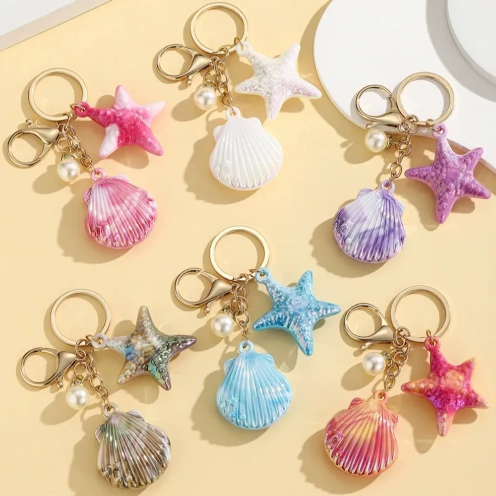 Six colorful resin ocean shell and starfish keychain charms with pearl beads in pink, white, purple, blue, iridescent, and hot pink, displayed on a yellow background.