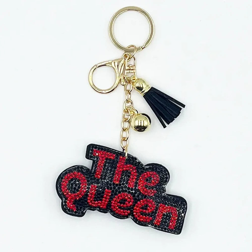 Two rhinestone-encrusted 'Bad Bitch' keychain charms in purple and pink with silver hardware, tassel, and lobster clasp, hanging on a cream bag. - Black