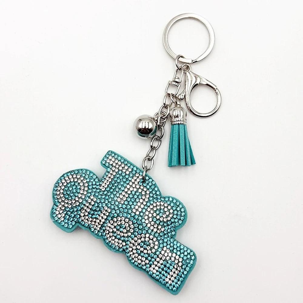 Two rhinestone-encrusted 'Bad Bitch' keychain charms in purple and pink with silver hardware, tassel, and lobster clasp, hanging on a cream bag. - Blue