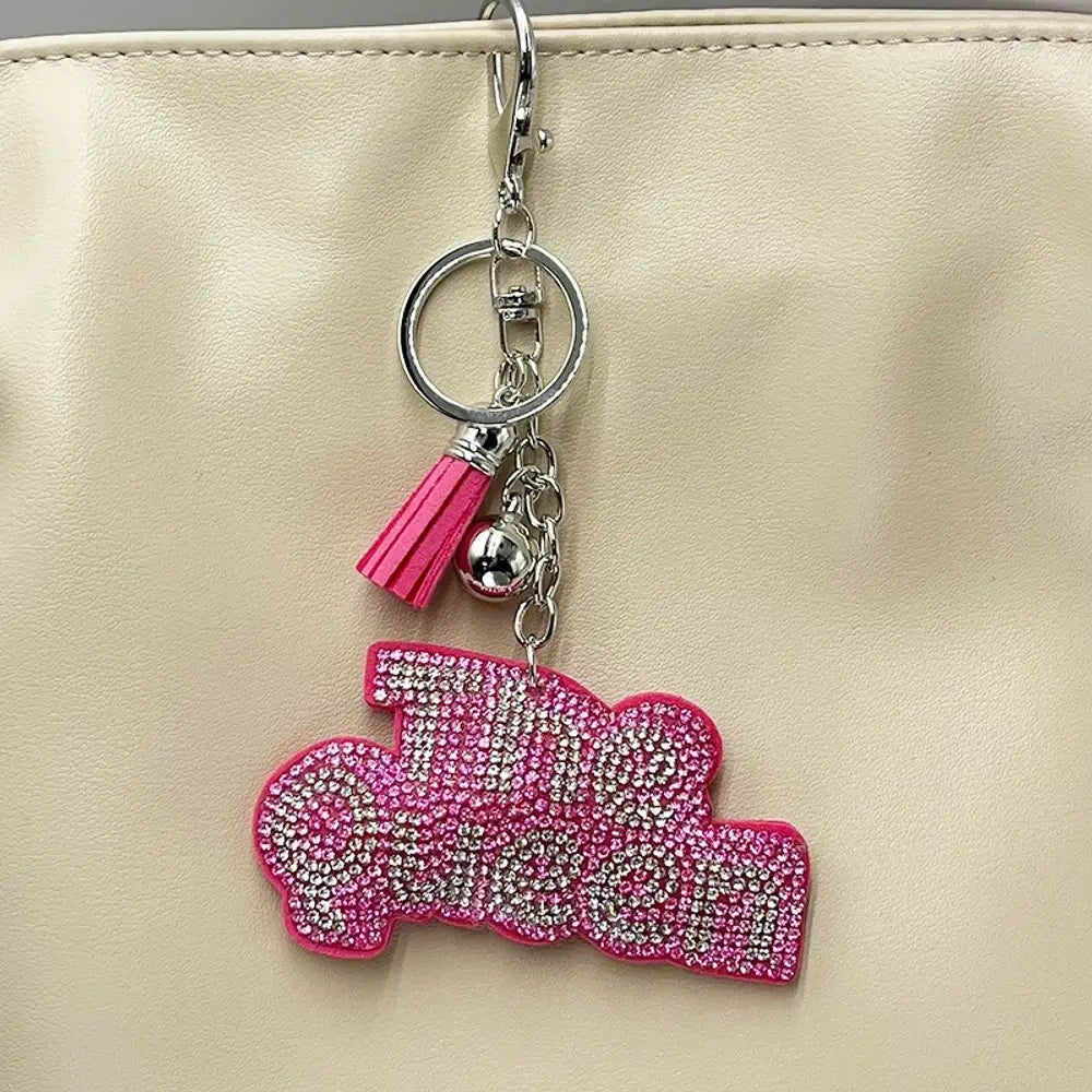 Two rhinestone-encrusted 'Bad Bitch' keychain charms in purple and pink with silver hardware, tassel, and lobster clasp, hanging on a cream bag. - Pink