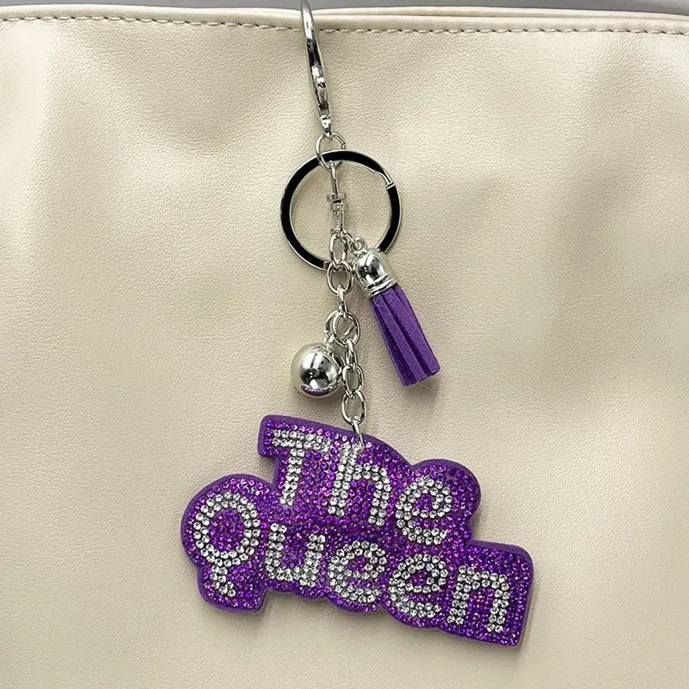 Two rhinestone-encrusted 'Bad Bitch' keychain charms in purple and pink with silver hardware, tassel, and lobster clasp, hanging on a cream bag. - Purple