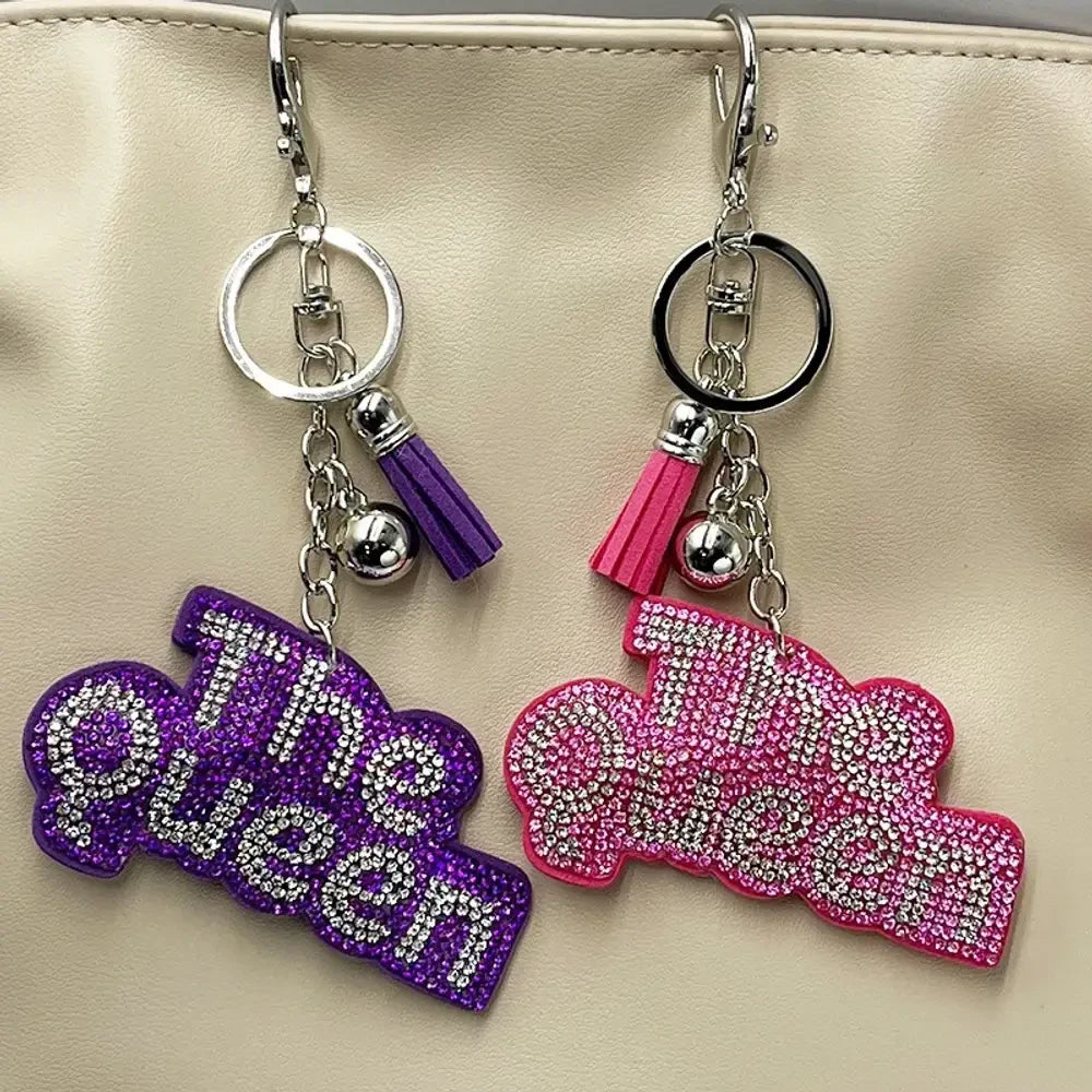 Two rhinestone-encrusted 'Bad Bitch' keychain charms in purple and pink with silver hardware, tassel, and lobster clasp, hanging on a cream bag. - Random Mixed