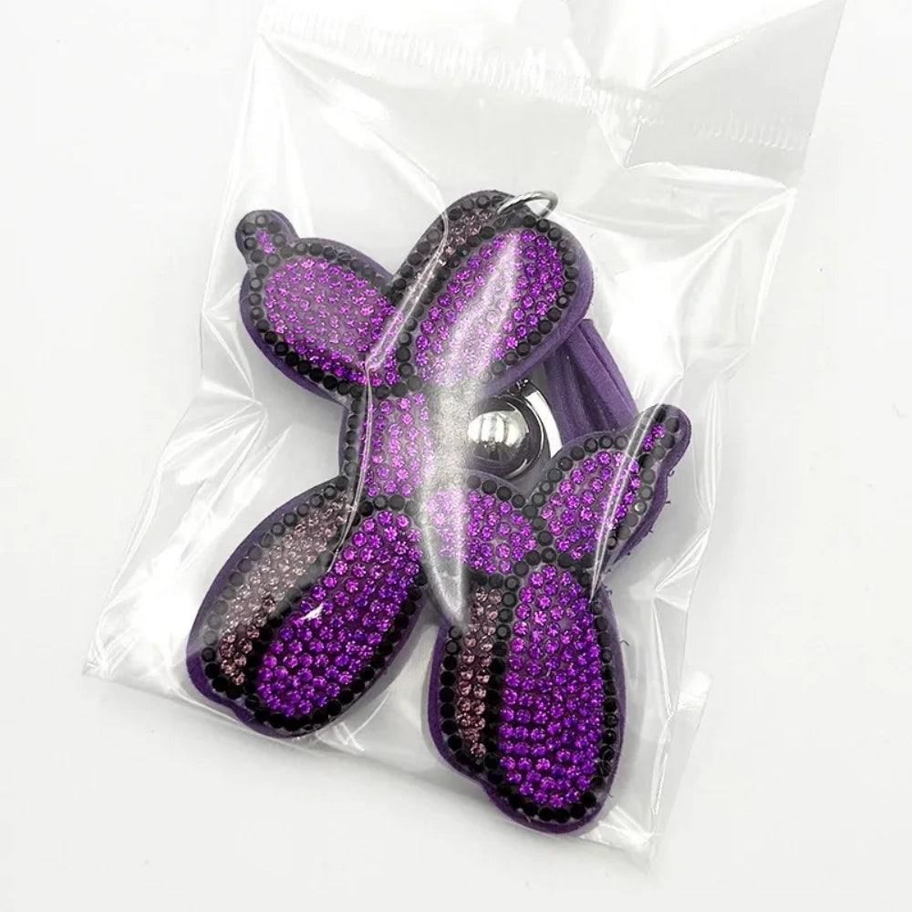 Five rhinestone-covered balloon dog keychain charms in green, yellow, brown, teal, and purple packaged in clear plastic bags on white background. - Purple