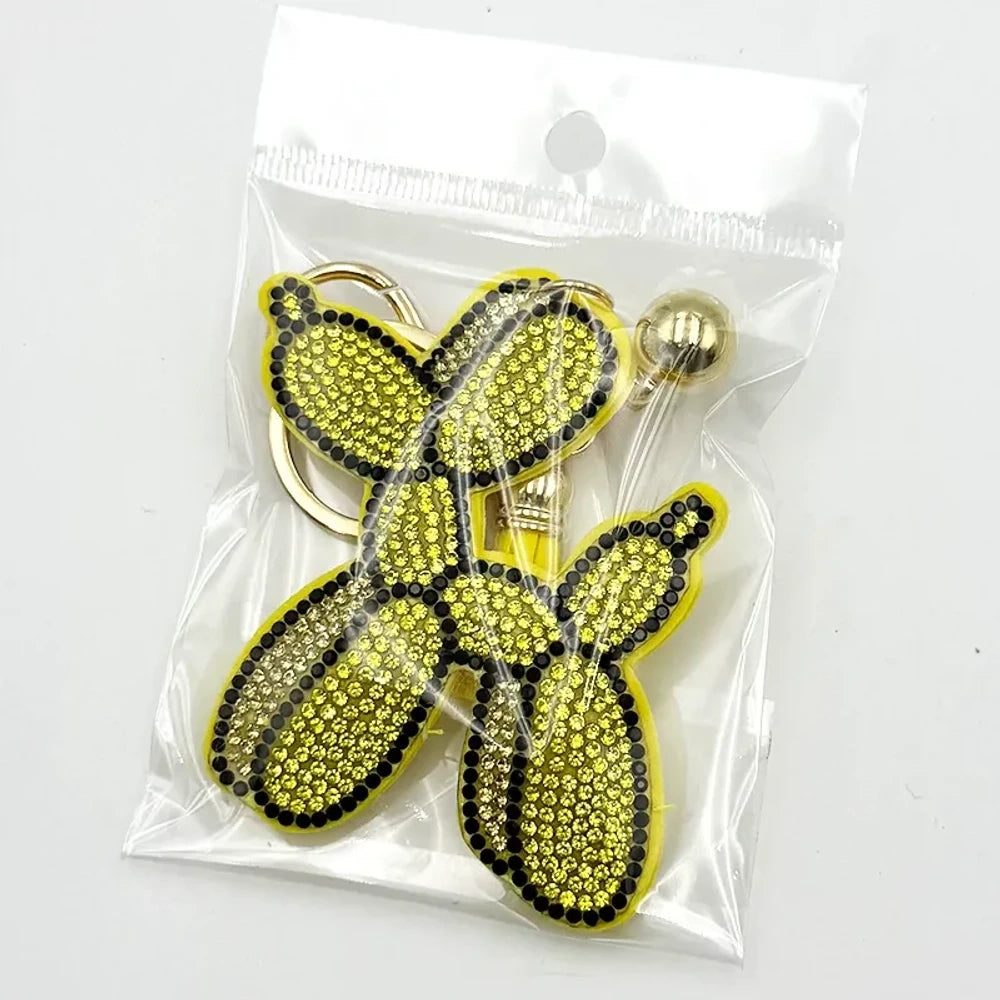 Five rhinestone-covered balloon dog keychain charms in green, yellow, brown, teal, and purple packaged in clear plastic bags on white background. - Yellow