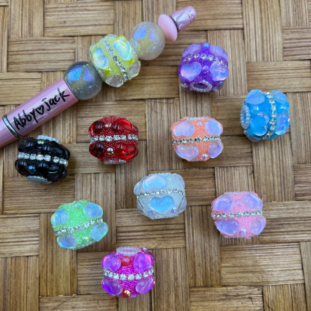 Assorted color rhinestone band bubble fancy beads in black, red, green, purple, pink, blue, peach, white, and yellow with opalescent finish displayed on woven rattan background next to a pink AbbyJack beadable pen.
