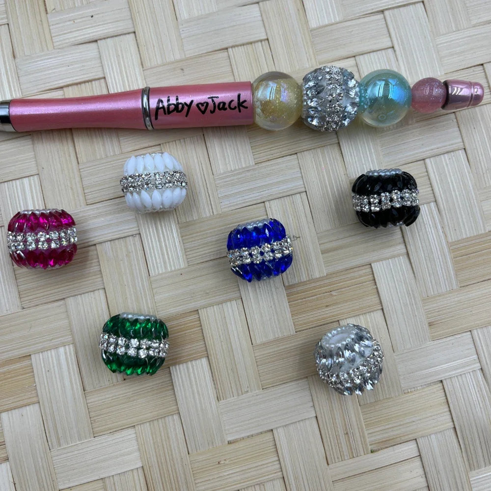 Assorted rhinestone banded crystal fancy beads in pink, white, blue, green, black, and clear displayed on a woven surface next to a beadable pen labeled Abby and Jack.