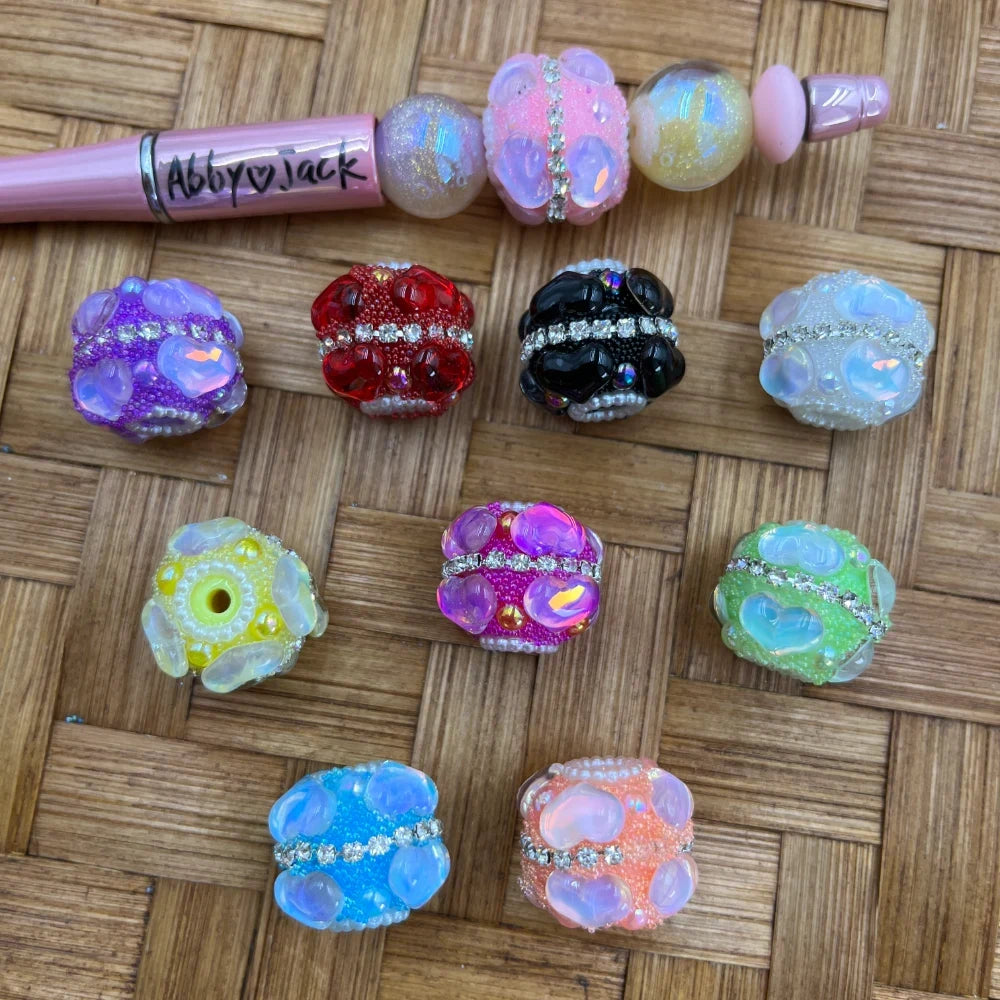 Assorted rhinestone studded berry cluster resin fancy beads in purple, red, black, white, yellow, magenta, green, blue, and orange displayed on a wicker surface alongside a pink beadable pen.