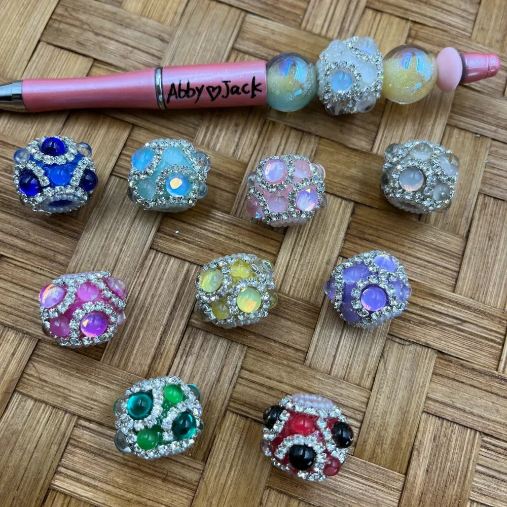 Assorted rhinestone studded bling fancy beads in blue, teal, pink, white, yellow, purple, green, and red with crystal embellishments, displayed on a woven surface next to a beadable pen labeled Abby and Jack.