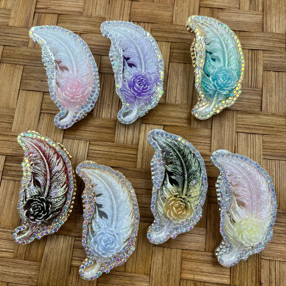 Seven rhinestone-bordered feather and rose resin flatback cabochons in pink, purple, teal, burgundy, white, black-gold, and ivory colors with iridescent AB crystal edges on a woven bamboo background.